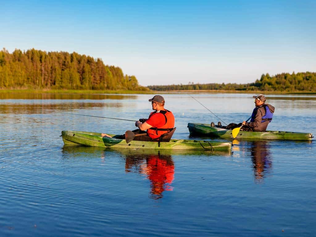 A side view of two anglers fishing from their kayaks on calm lake waters against fall scenery in the background