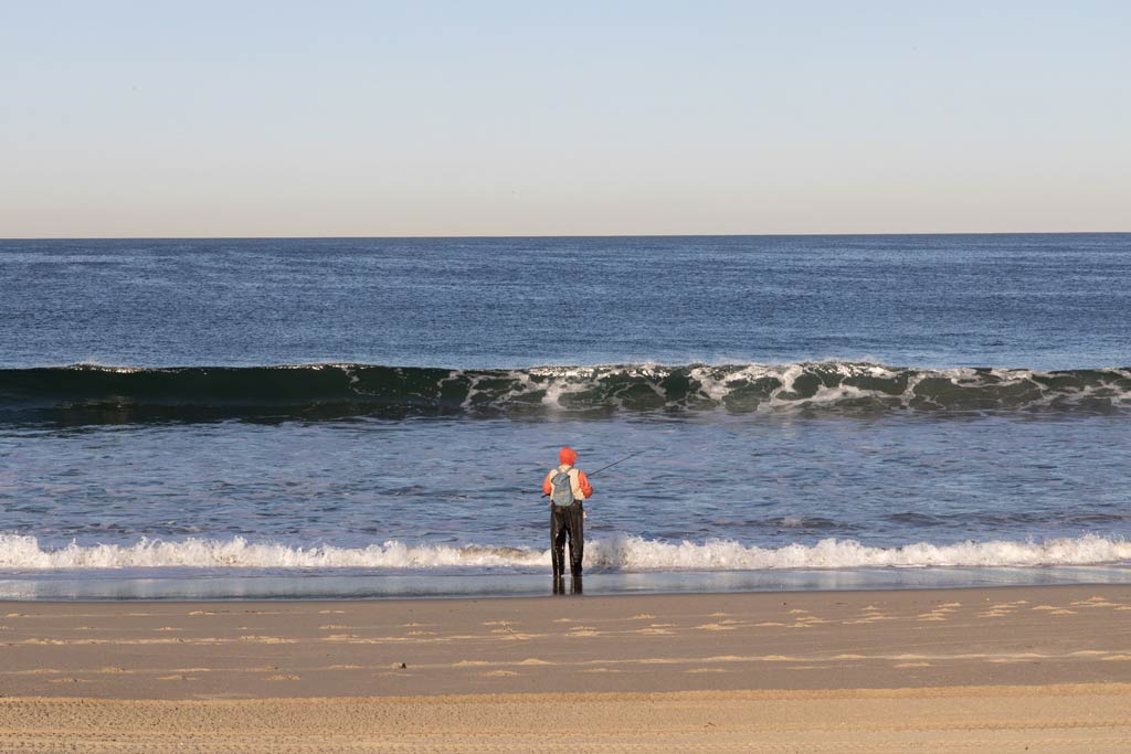A lone angler standing in the surf and fishing at Redondo Beach, as a wave crashes in on a clear day
