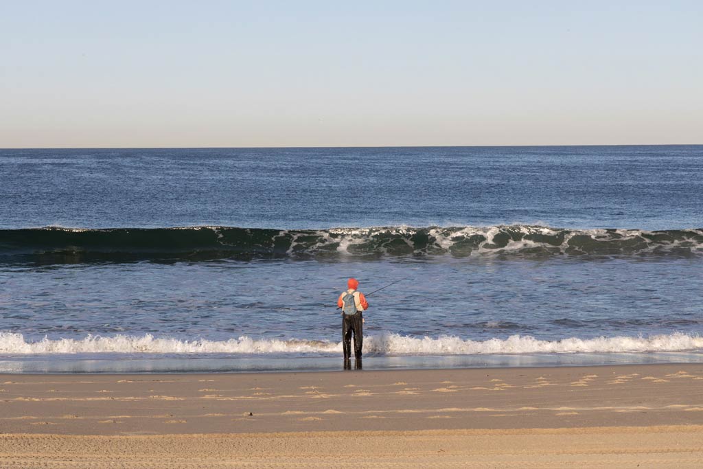 A lone angler standing in the surf and fishing at Redondo Beach, as a wave crashes in on a clear day