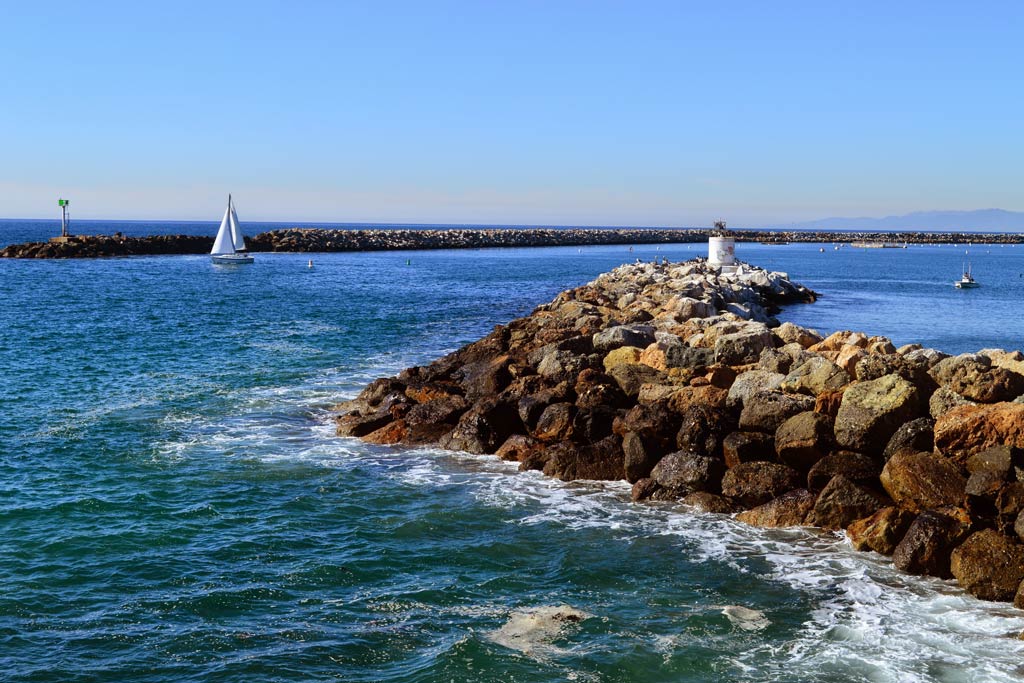 A rocky Redondo Beach jetty with blue water surrounding it and blue skies beyond, along with a couple of smaller boats 