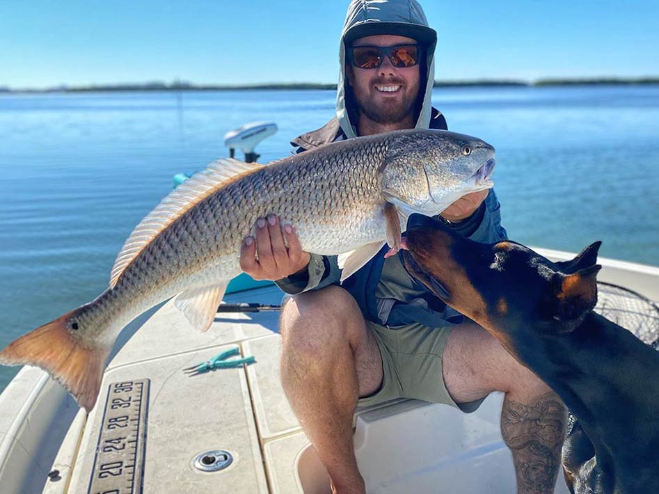 An angler sitting on a boat and holding a Redfish he caught while his dog curiously approaches it, Tarpon Springs, Florida