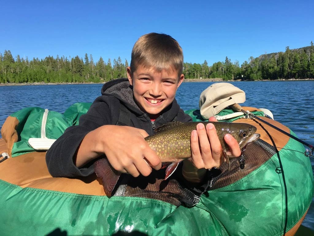 A photo of a kid smiling while holding a small Brook Trout with both hands and posing for the camera on a green kayak somewhere in Michigan.