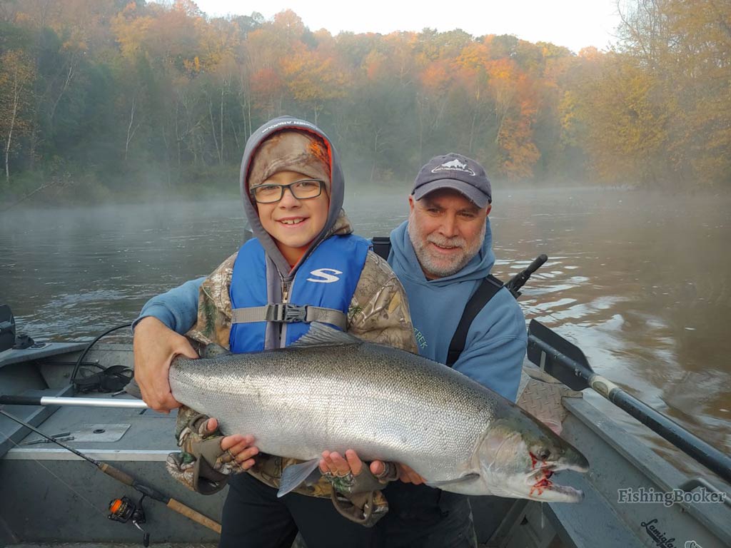 A child and an elderly man sitting on a fishing boat in Michigan, holding a Coho Salmon, with a misty river and autumn foliage in the background