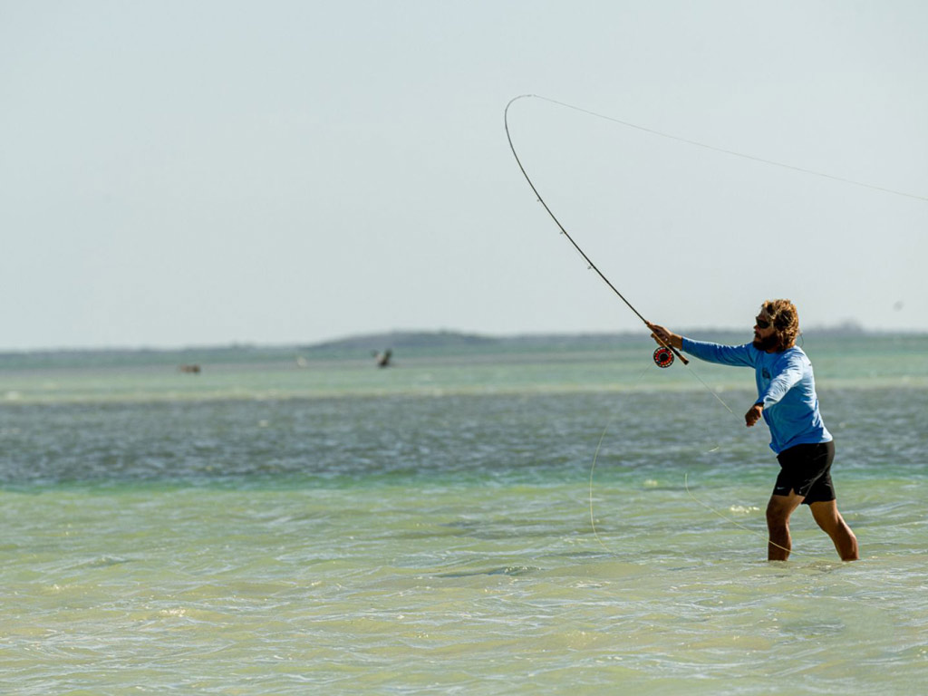 A man with long hair and a beard standing in ankle deep water in the flats in the Florida Keys, casting with his fly rod high up in the air.