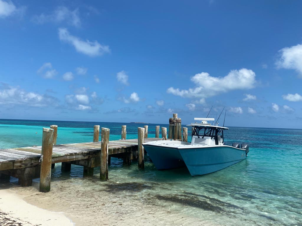 A catamaran fishing boat next to a wooden dock on a beach, with beautiful blue waters and clear skies around it.