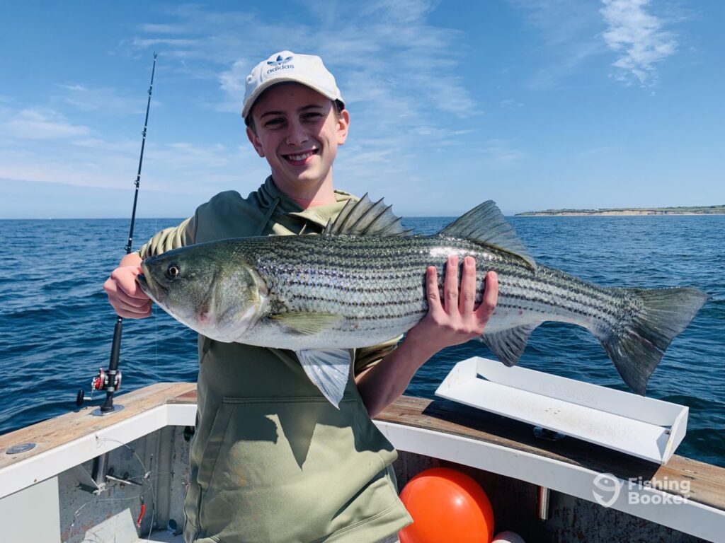 A person proudly holding a large Striped Bass on a boat, showcasing their angling skills in one of the best US fishing states, with gear visible nearby.