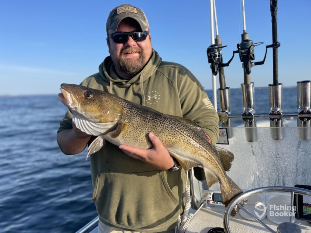 A person proudly displays a large Cod on a boat in one of the best US fishing states, sporting sunglasses and a cap, with fishing rods ready in the background.