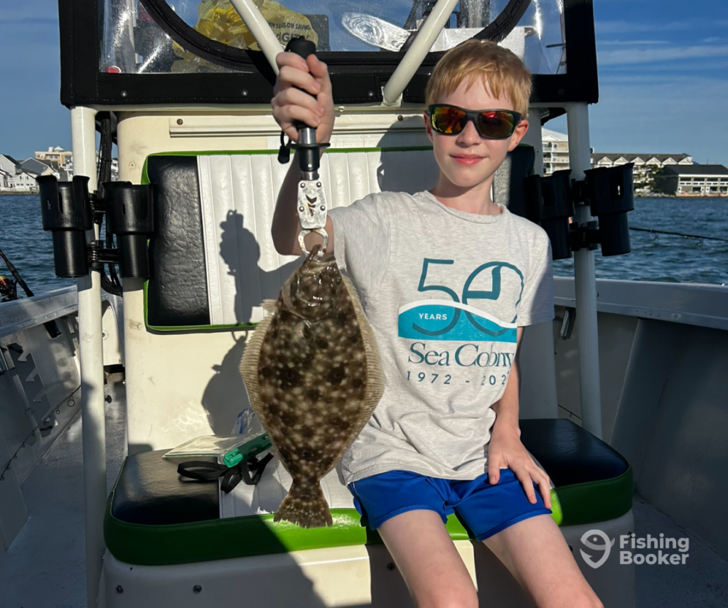 A young person in sunglasses, holding a Flatfish on a boat, while sitting on a bench. With water and buildings visible, they're enjoying one of the best US fishing states.