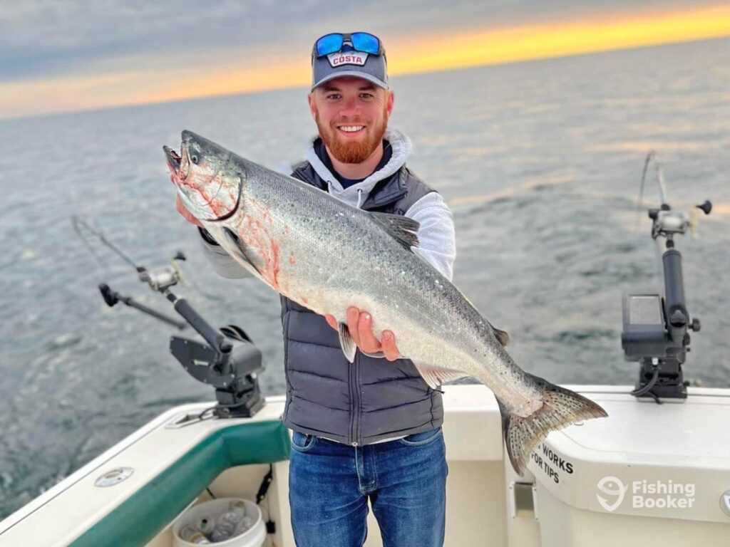 A man proudly holds a large Salmon on a boat, showcasing the bounty of one of the best US fishing states, with the ocean and sunset painting a stunning backdrop.