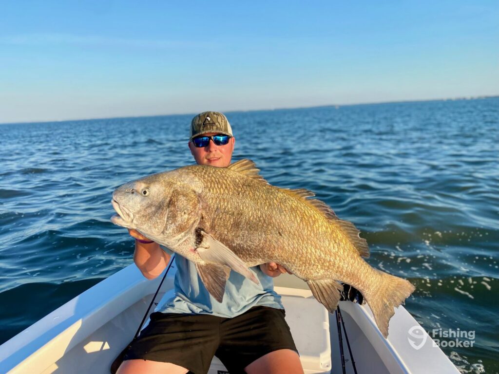 A person proudly holds a large Drum fish on a boat in open water, wearing sunglasses and a cap, embodying the thrill of fishing in one of the best US fishing states.