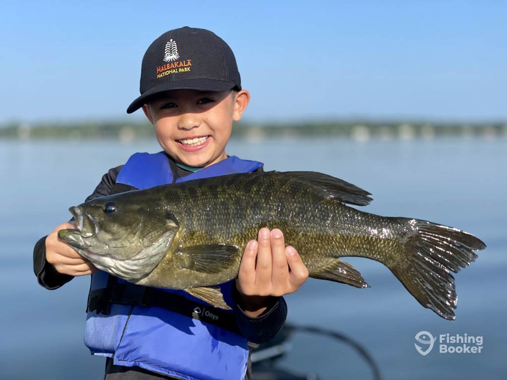 A child in a blue life jacket proudly holds a large Smallmouth Bass with both hands while on a boat, enjoying Fox River fishing. The calm water reflects the sunny sky, and you can see a distant shoreline in the background.