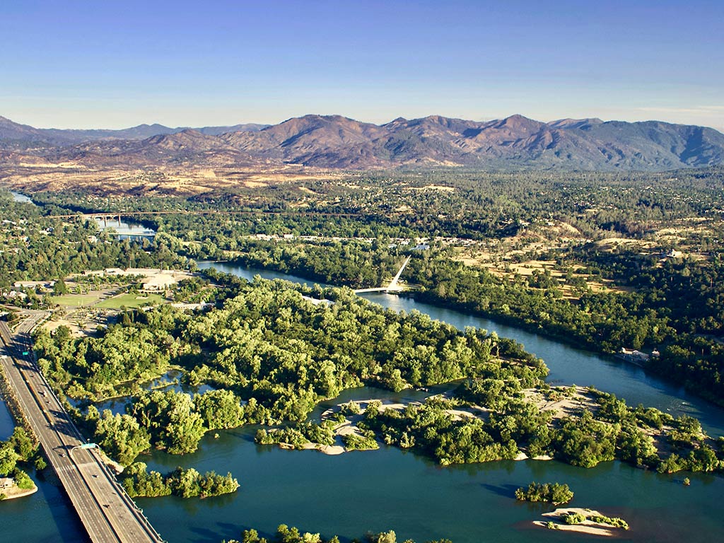 An aerial view of Redding, California, taken on a clear, sunny day, showing the greenery that surrounds the town and the river that runs through it, with some hills in the distance