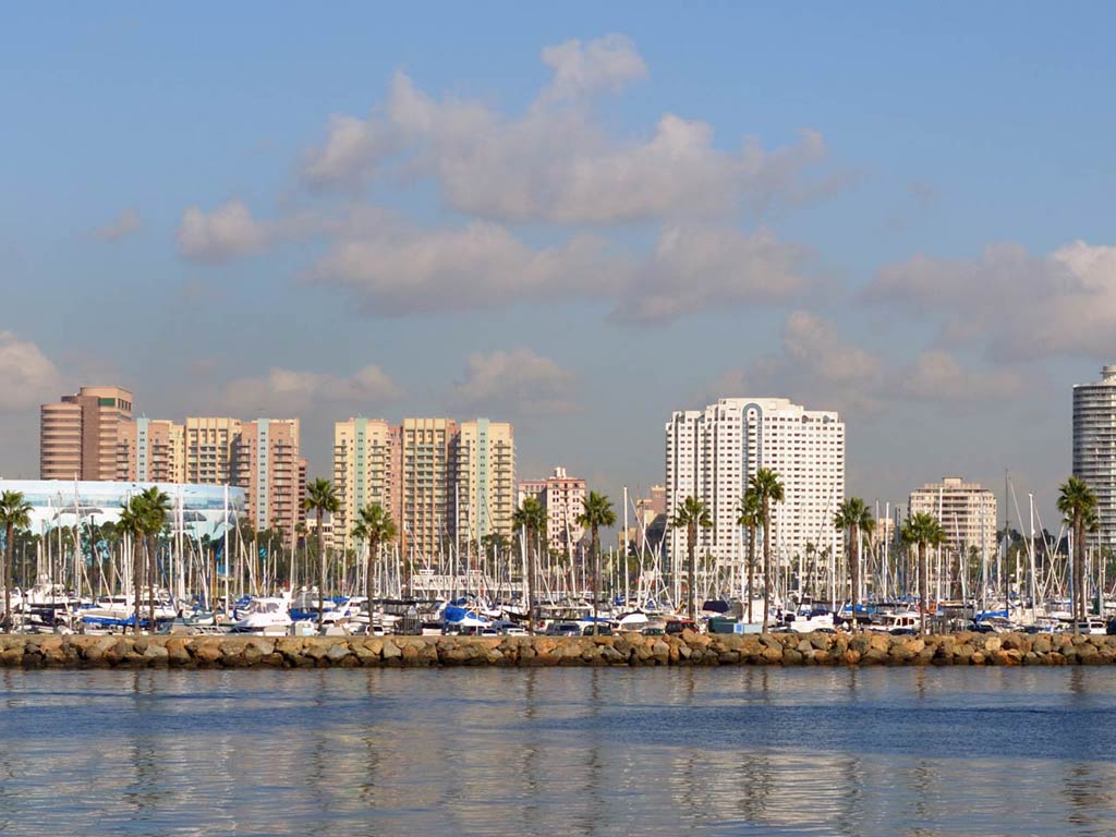 A photo of Long Beach taken from the water so that you can see the city’s skyline, its buildings, the palm trees, and boats on a bright day.