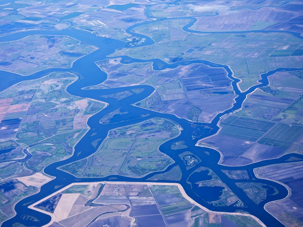 An aerial view of the meandering California Delta and its surrounding greenery and fields as seen on a bright day