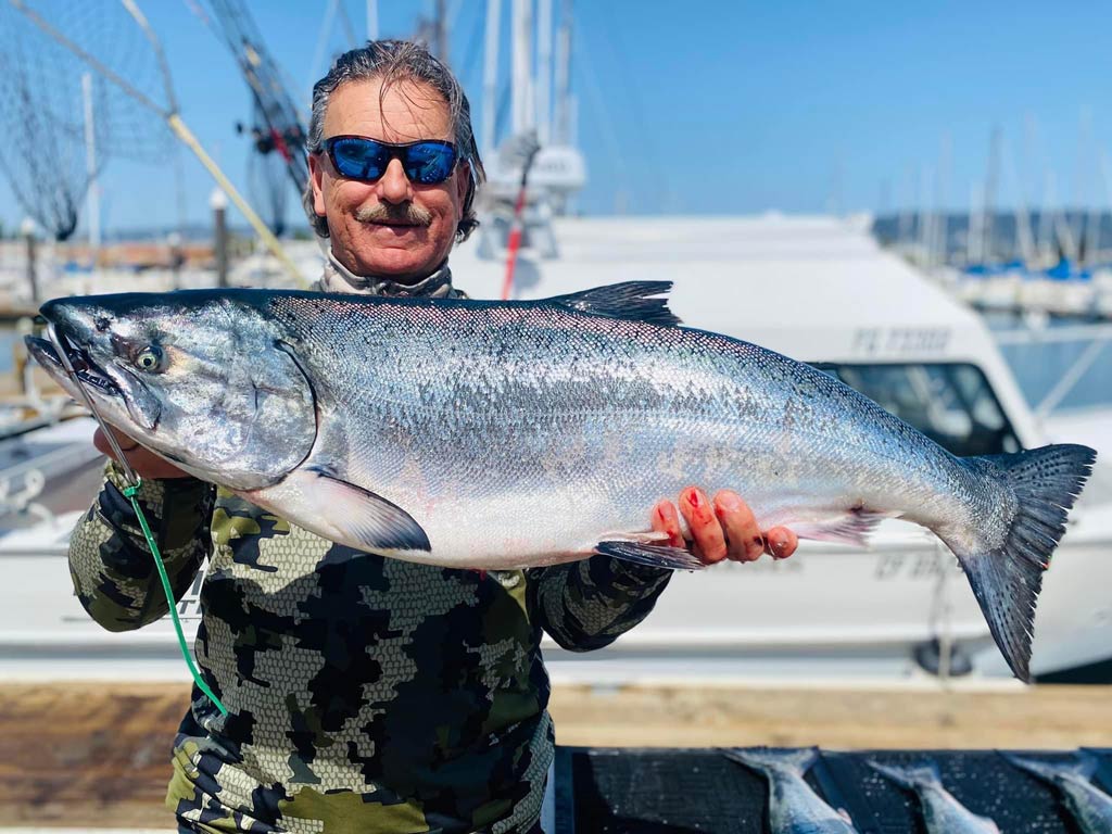 A photo of an angler wearing a pair of sunglasses while posing on the dock in front of charter boats with a big Chinook Salmon in his hands.