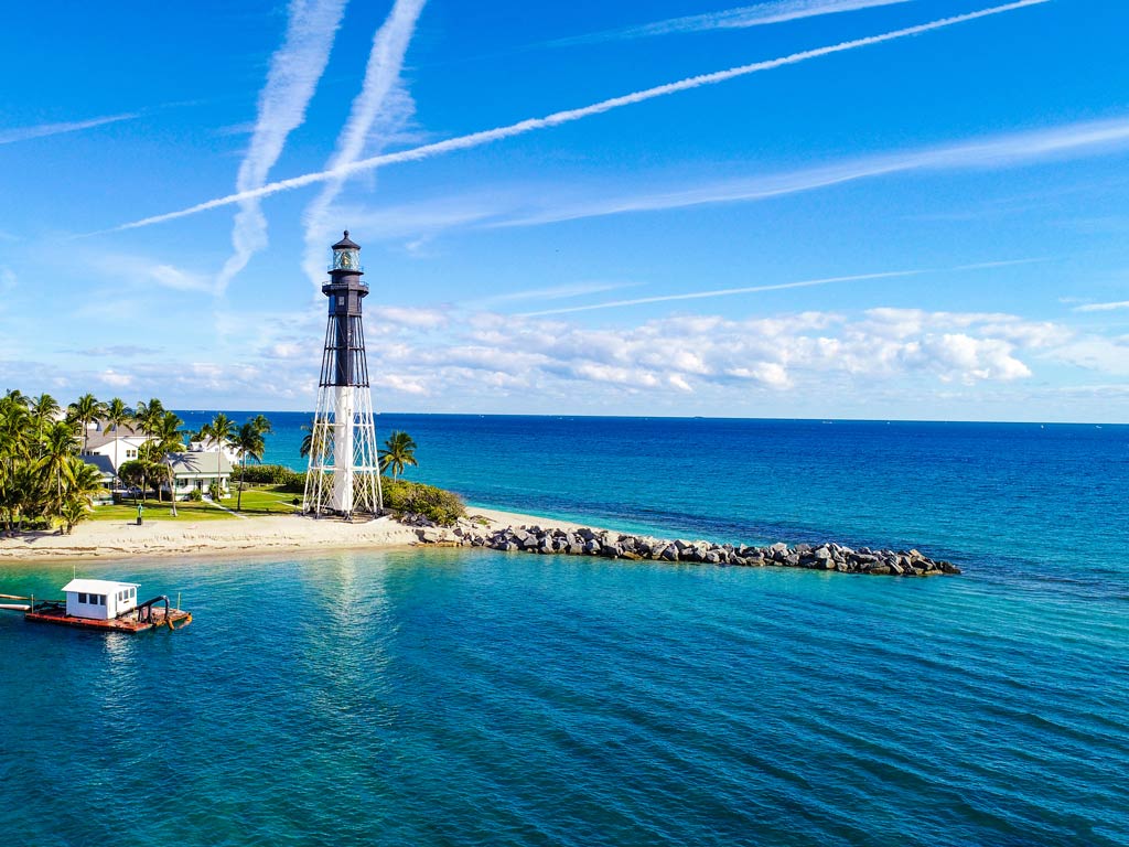 A view of the Hillsboro Inlet Lighthouse, with a jetty in front of it, a sandy beach and clear waters surrounding it, and blue skies in the background