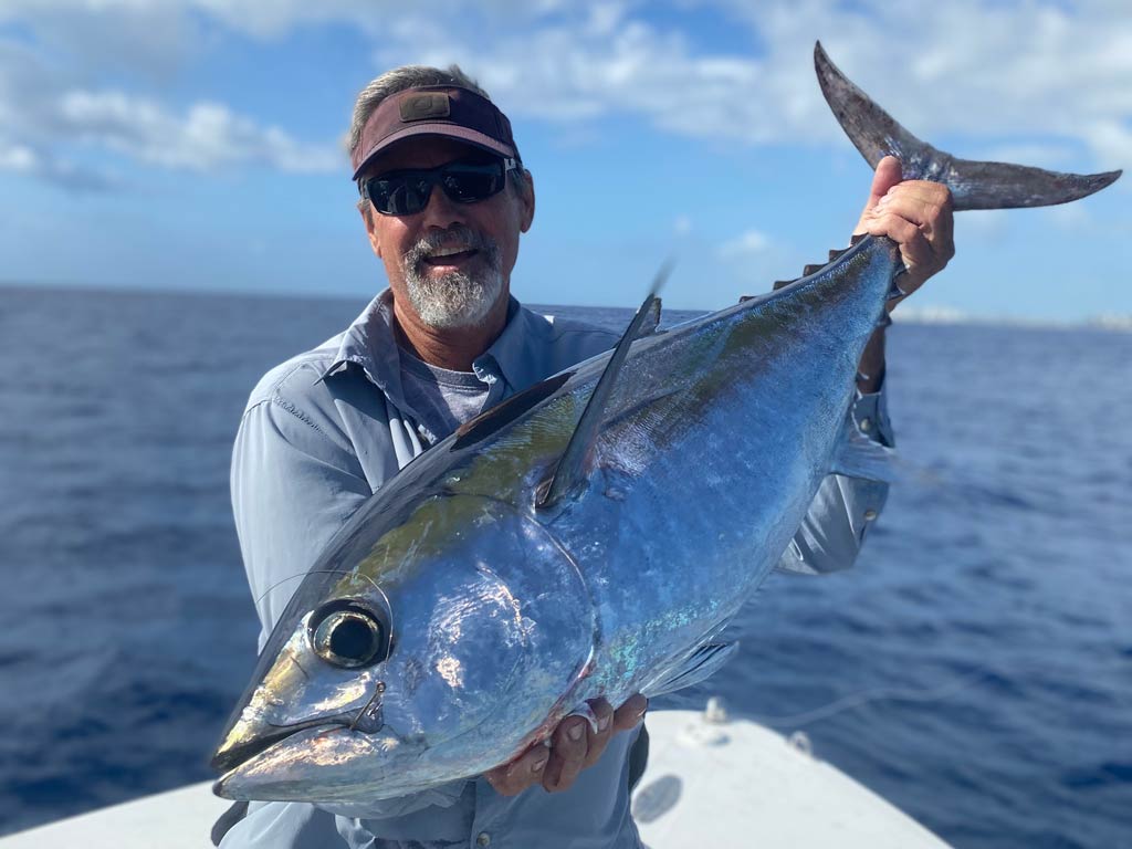 A smiling middle-aged man in a cap and sunglasses standing on a charter boat, holding a Blackfin Tuna, with blue skies and open waters in the background