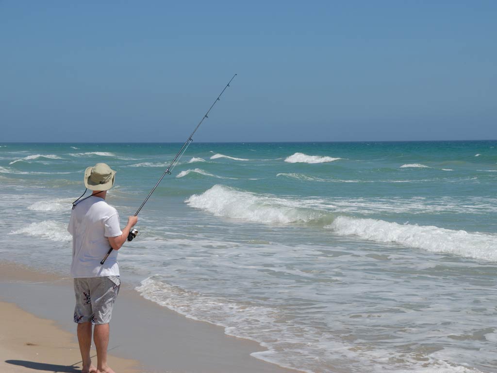 A fisherman standing on a sandy beach holding on a fishing rod and looking away from the camera, with a soft surf coming in