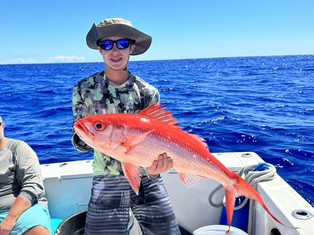 A young angler proudly holding Onaga Long Tail Red Snapper while standing on a Jigging Honolulu charter fishing boat