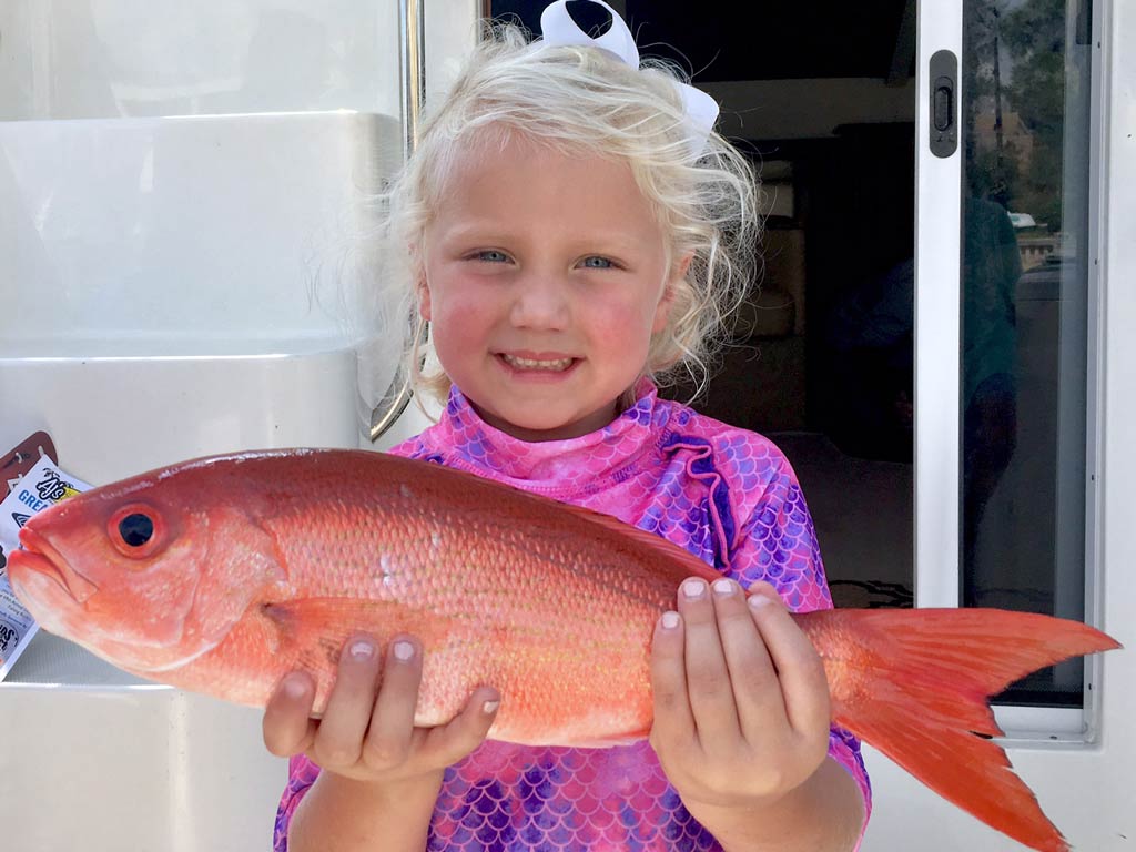 A little girl smiling while standing on the dock in front of a charter boat and posing with Vermillion Snapper in both hands.