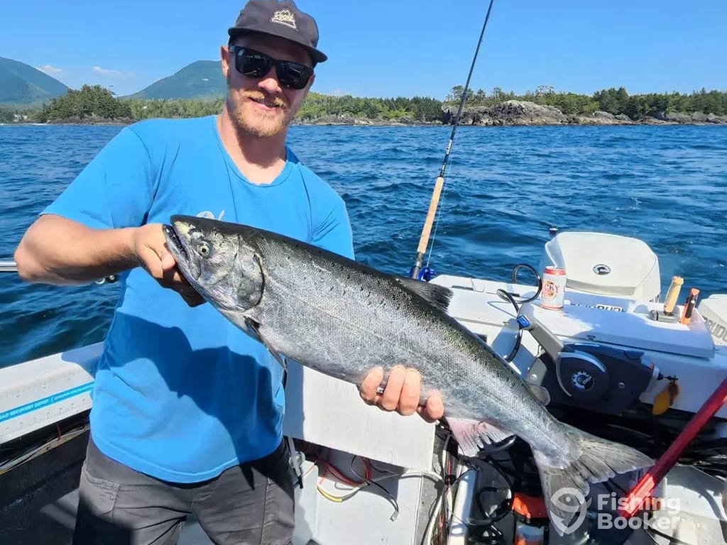 An angler in sunglasses, a baseball cap, and blue t-shirt holds a sizeable Coho Salmon aboard a fishing boat with the water behind him on a sunny day