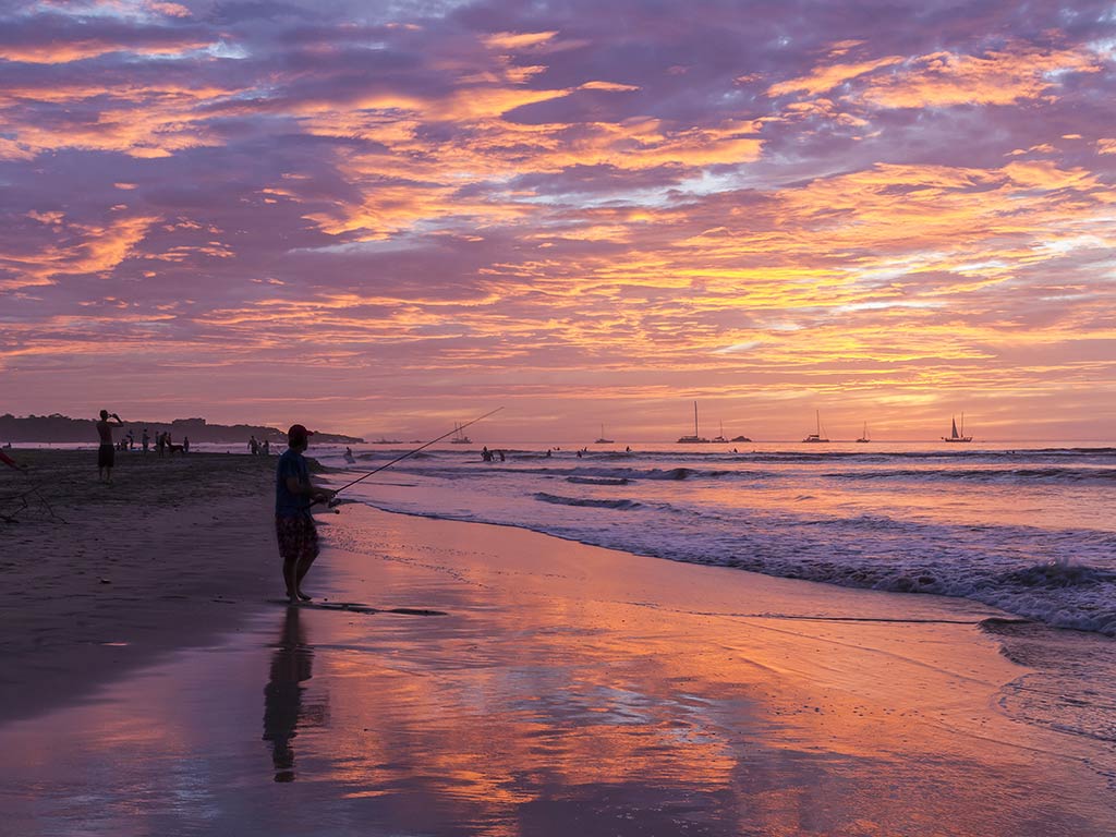 A view along a beach in Tamarindo at sunset, with a lone angler casting a fishing line into the Pacific Ocean
