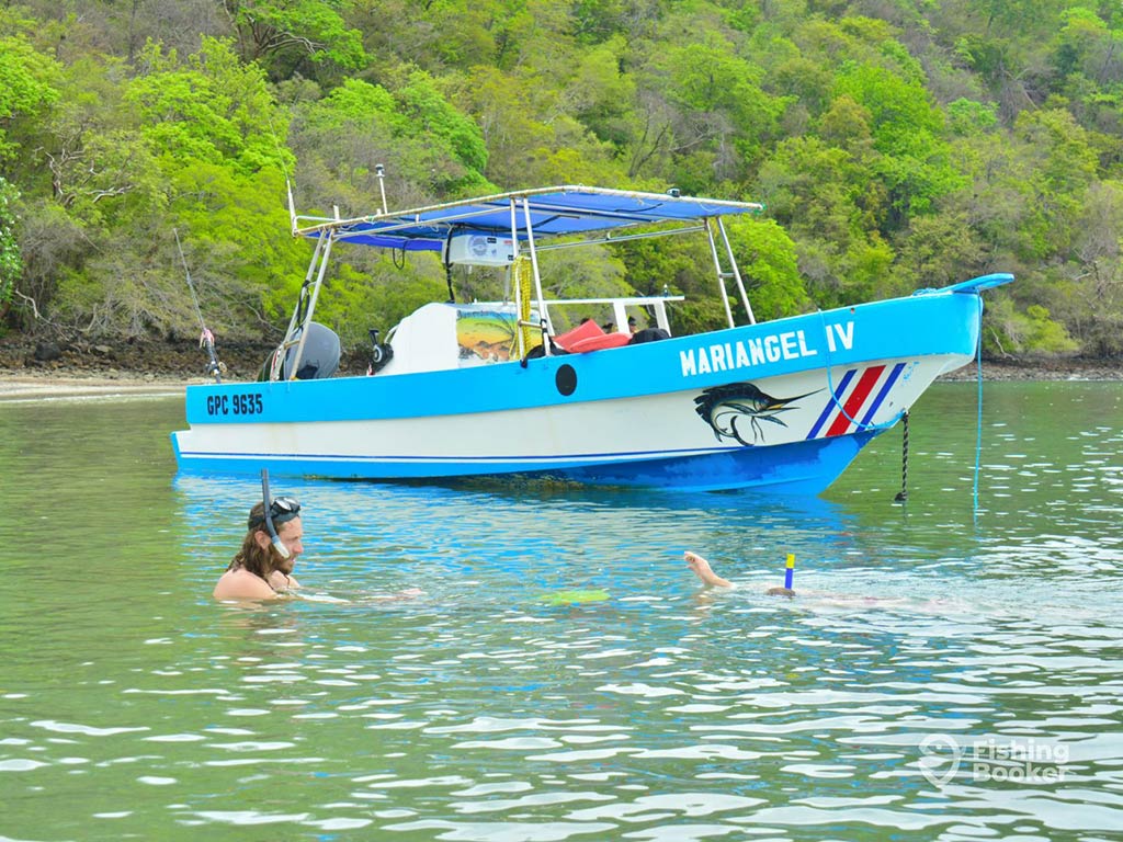 A modern, blue panga fishing boat docked in the shallow waters of Tamarindo, with a snorkeler relaxing in the water in the foreground and some green foliage in the distance