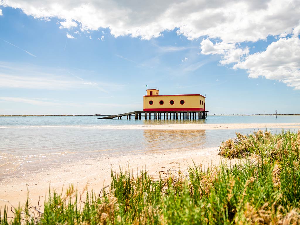 A view from shore towards the historic Rio Formosa lifeguard station, abandoned in the water on a day with sunny intervals, with the beach visible in the foreground