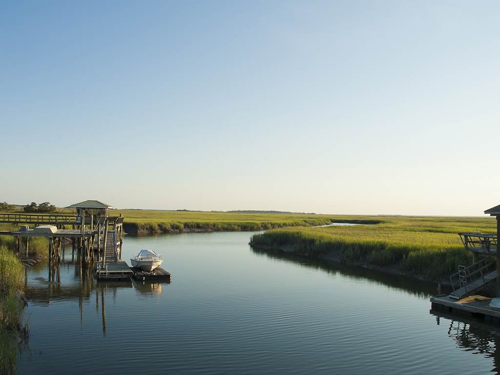 A view across a winding waterway in the backcountry around Savannah, with a small, wooden fishing pier and boat dock visible on the right of the image at sunset and grassy flats on either side of the water