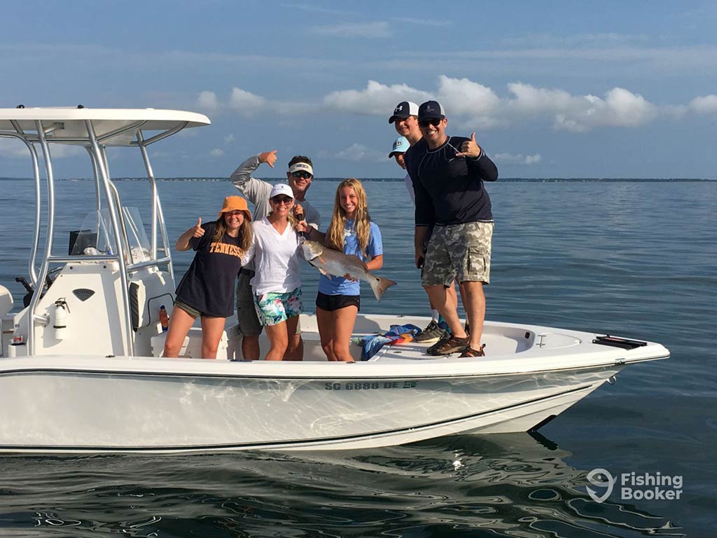 A view across the water towards a center console charter boat with a group of anglers posing for the camera and holding up one Redfish on a sunny day