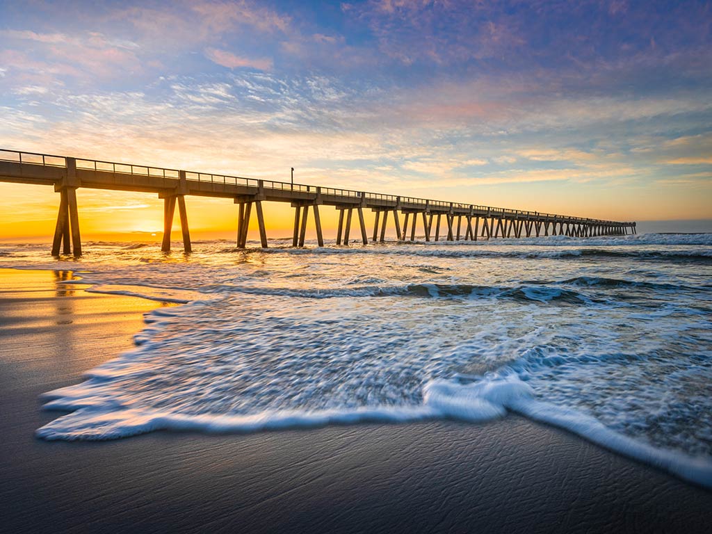 A view from the beach of the lengthy fishing pier on Navarre Beach, with the water crashing into the beach at sunset