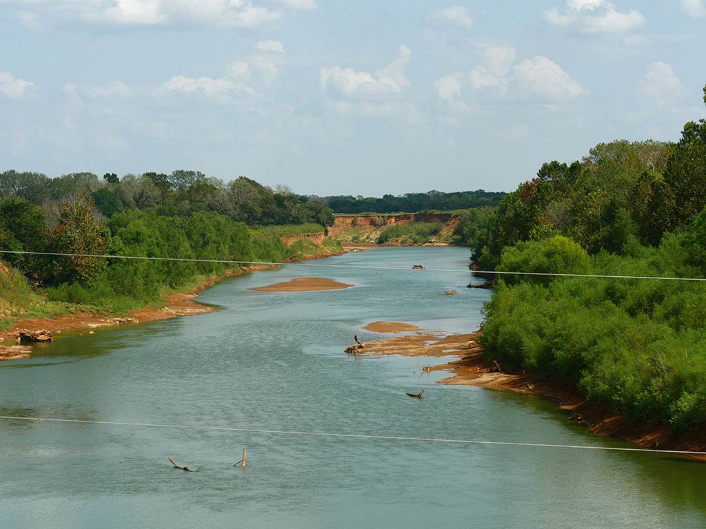 An aerial view looking down the Washita River on a cloudy day, with green trees lining either bank and telephone lines cutting across the center of the image