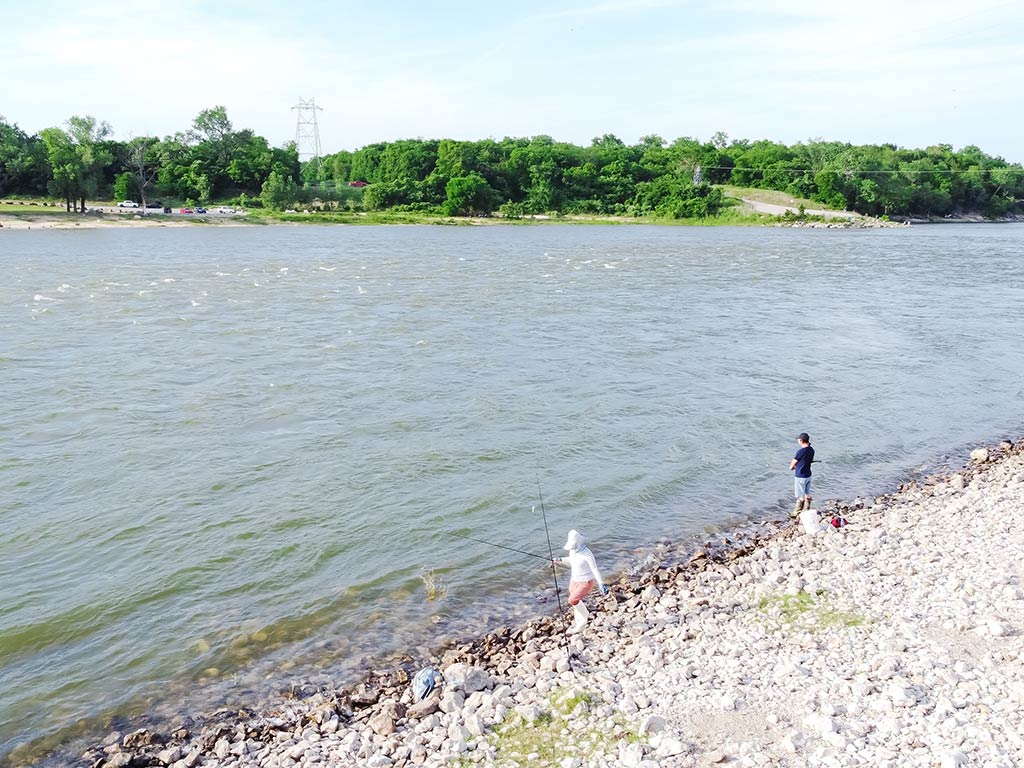 A view from a hill towards two anglers shore fishing along the Red River in Oklahoma, with green trees visible on the opposite shoreline on a cloudy day
