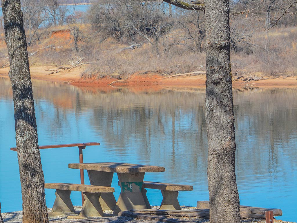A view of a concrete table and benches in a picnic area in Kingston, OK, with Lake Texoma Visible through the trees in the distance on a sunny day