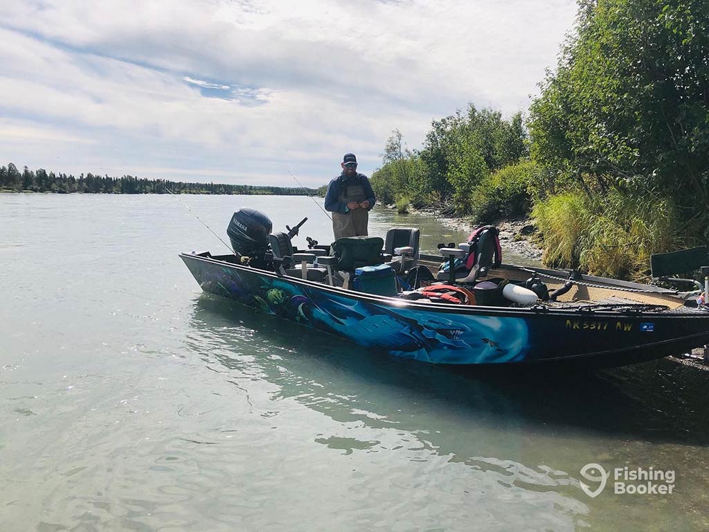 A man stands on a fishing boat on a river in Alaska, setting up his fishing gear, with the calm waters and a green landscape surround him on a day with sunny intervals