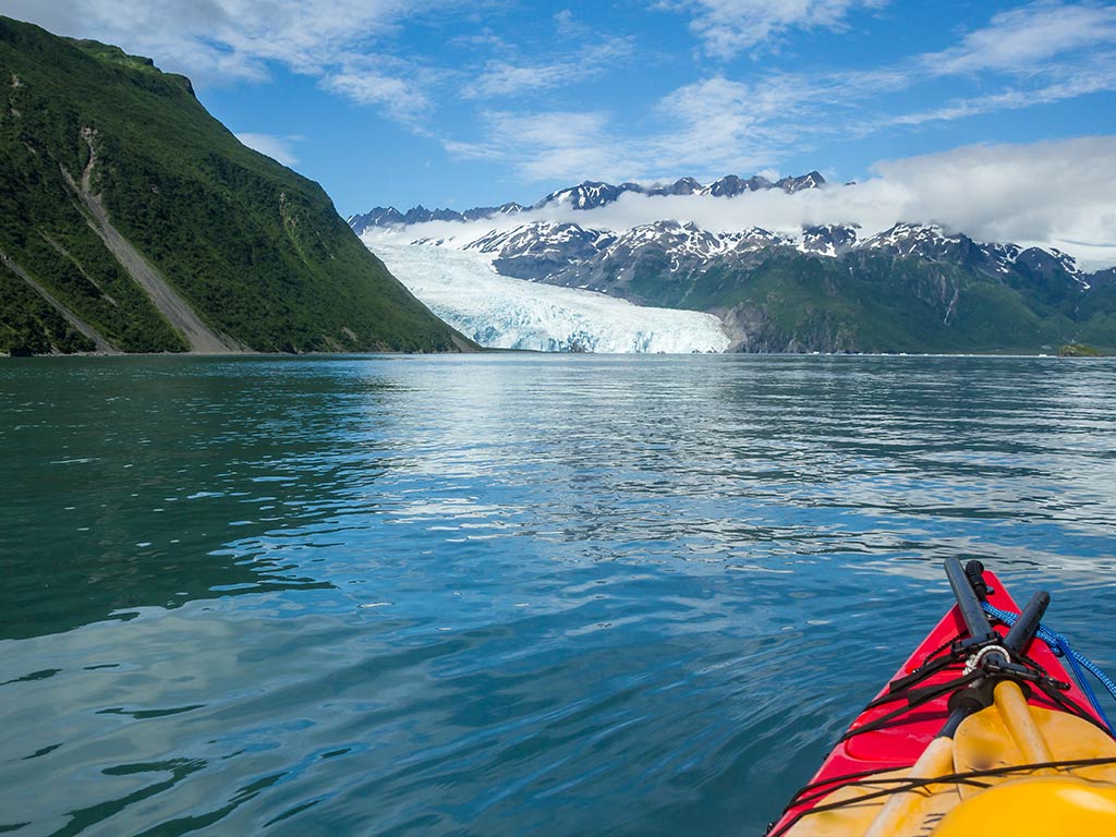 A view from a kayak with the tip of it visible in the foreground towards a glacier in Kenai Fjords National Park on a day with sunny intervals
