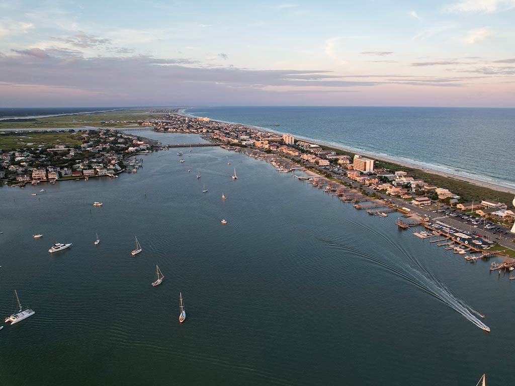 An aerial view of Wrightsville Beach, NC, with an inshore bay visible in the foreground, giving way to a barrier island full of houses and the ocean in the distance
