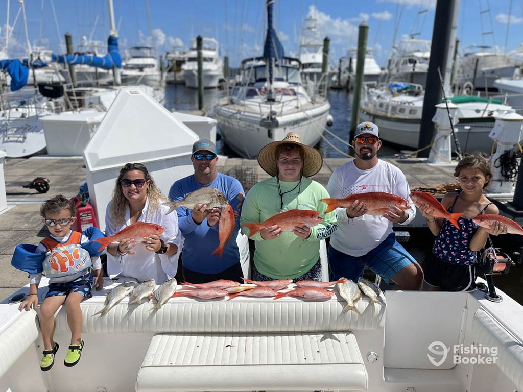 A family of anglers stood and at on the dock behind the deck of their fishing boat, presenting their catch from the day – mainly Snappers – by holding them and with some laid out in front of them on the back of the boat