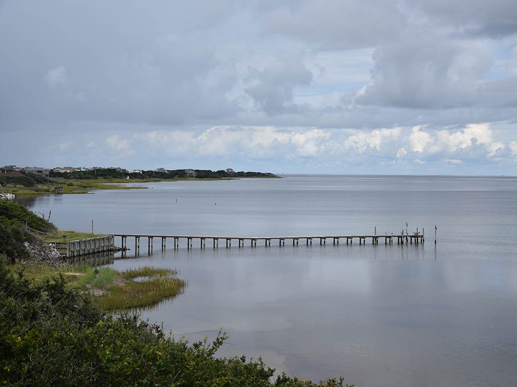 A view from a hill towards the Pamlico Sound on a cloudy day, with a small, wooden fishing pier sticking out into the water in the middle of the image