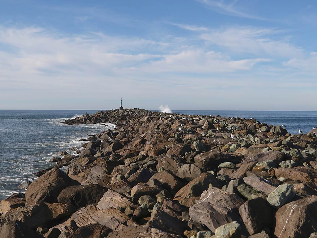 A view from the shore along a rocky jetty towards a distant lighthouse in Garibaldi, OR, on a day with sunny intervals, with the waves crashing into the rocks from the left