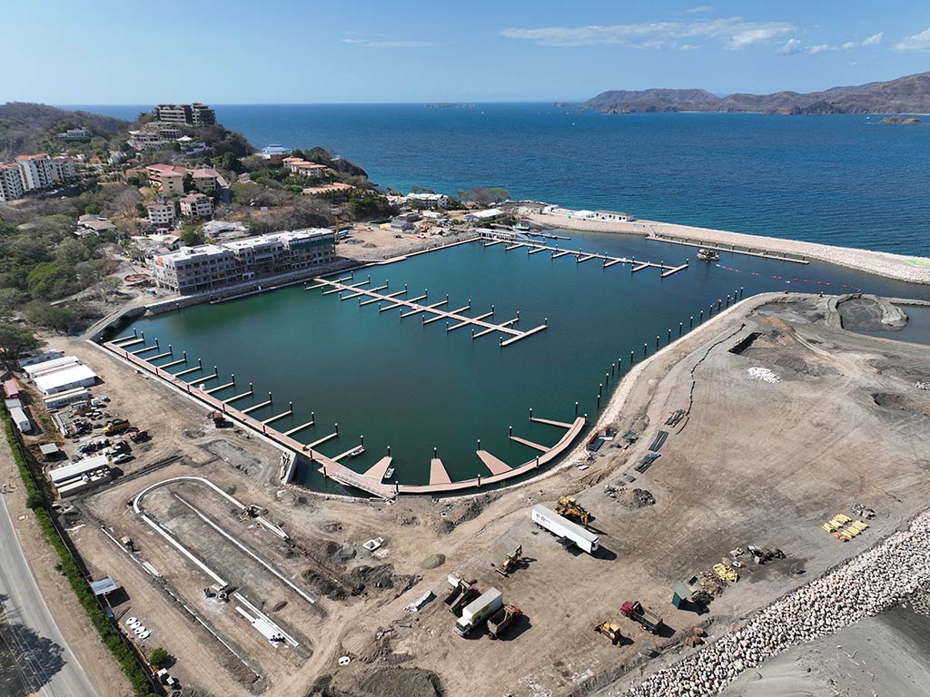 An aerial view of a marina in Playa Flamingo, Costa Rica, on a sunny day, with a concrete road visible in the foreground and the ocean in the distance