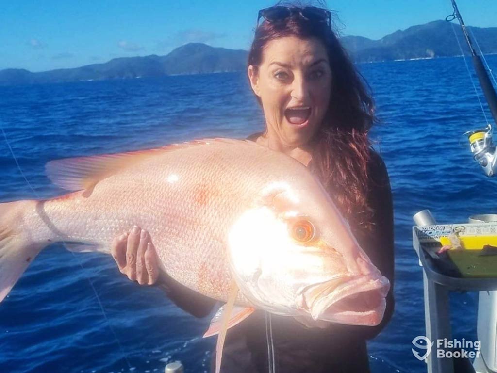 A woman with sunglasses on her head, looking surprised as she holds a large Nannygai fish aboard a charter in Airlie Beach on a sunny day