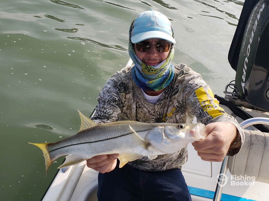 An angler in a light blue baseball cap and matching neck bluff crouching on a fishing charter in the Laguna Madre, holding a Snook with the water visible behind him