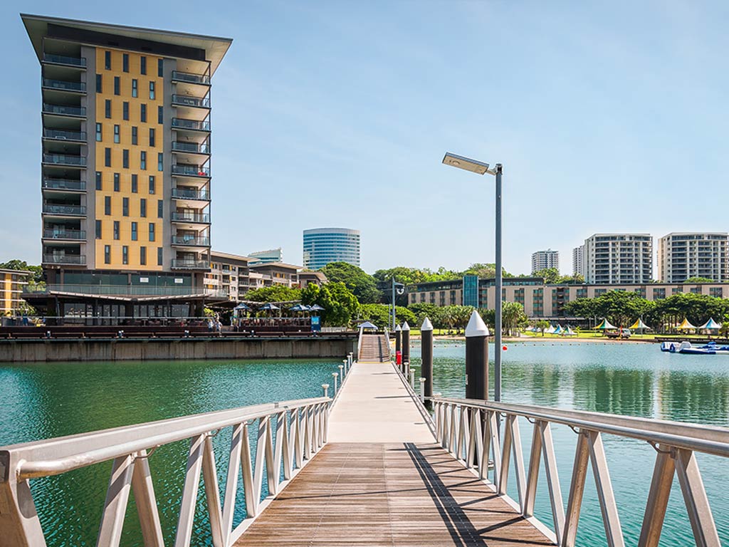 A view along a wooden dock towards downtown Darwin, Australia, on a clear day, with a large building on the left of the image and clear waters visible around the dock