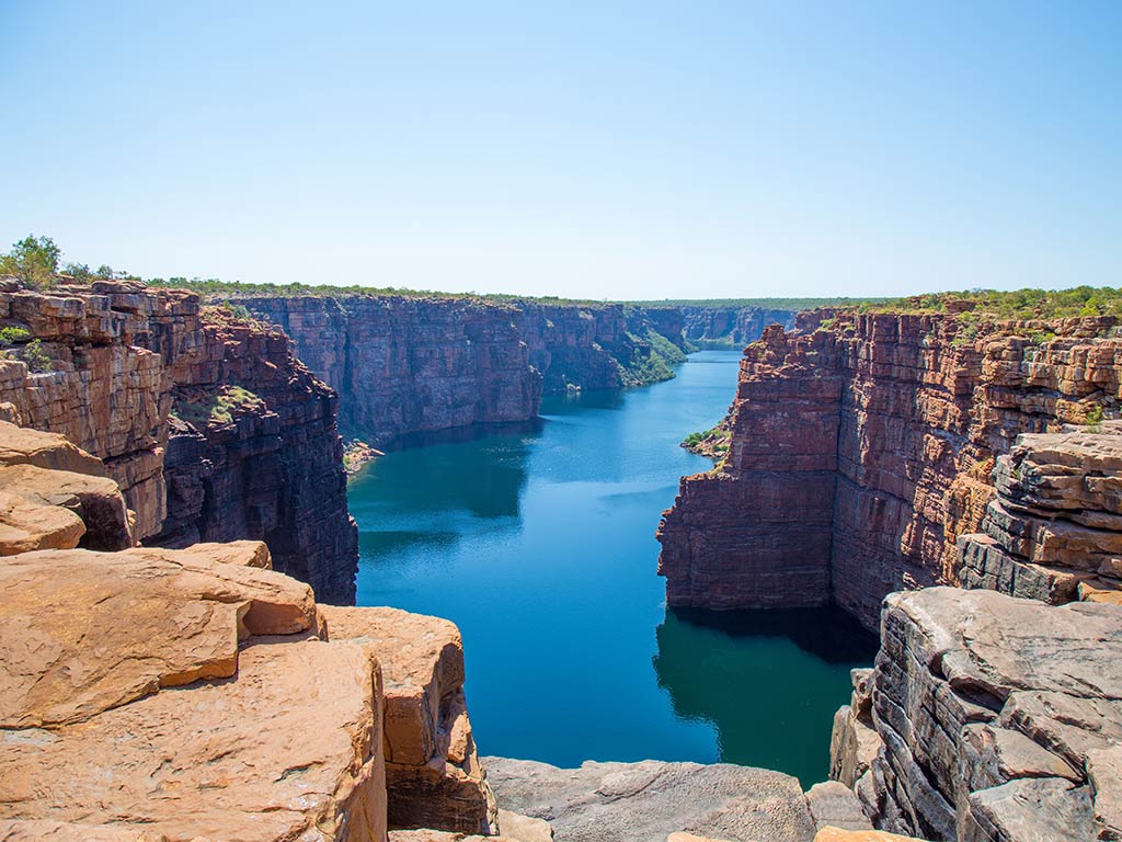 A view from the top of a cliff towards a stunning gorge near Darwin, Australia, on a clear day, with red rocks towering either side of pristine blue waters