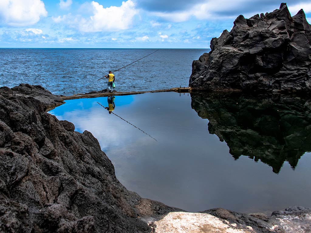 A view from behind of an angler walking into the water in Madeira and casting his line in between rocks