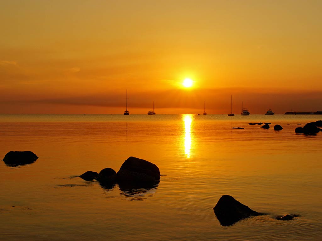 A view out from a beach in Tasmania to the sun setting in the distance, creating an orange hue over calm waters