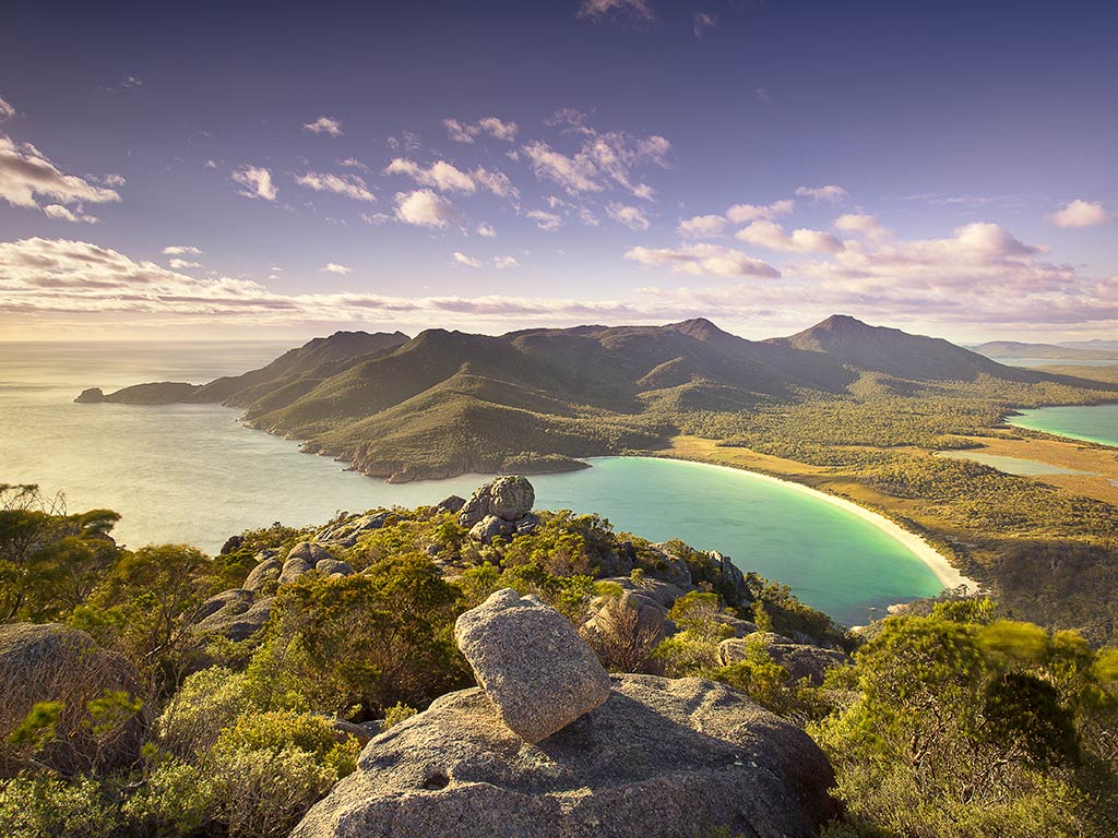 An aerial view from Mount Amos, Tasmania, towards the crystal clear waters of a bay leading out into the ocean on a sunny day