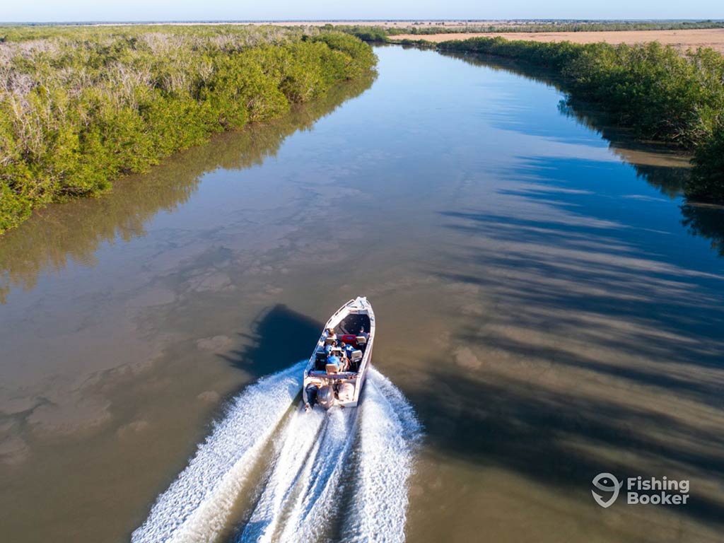 An aerial view of a charter fishing boat cruising along a brackish river in the Northern Territory, flanked by two tree-lined shores, leaving a noticeable wake behind it