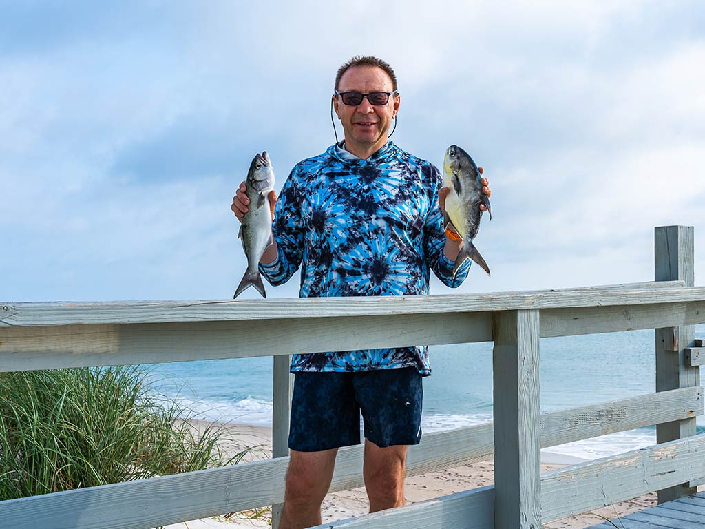 A person in a patterned shirt is holding two fish, standing behind a wooden railing with the beach and ocean visible behind him in Florida.
